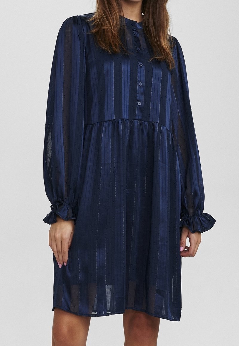 Granatowa sukienka wykonana z przejrzystej, fakturowanej tkaniny z pionowymi paskami i marszczonymi taliami. Posiada zapięcie na guziki z przodu oraz bufiaste rękawy.