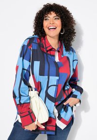 Camicia a maniche corte multicolore con motivi geometrici blu, rossi e neri, dal design con colletto e vestibilità rilassata. Borsa bianca tenuta in mano.