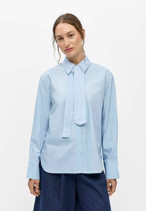 Young woman with light brown hair wears a light blue long-sleeve shirt with a fabric tie detail and dark blue wide pants.