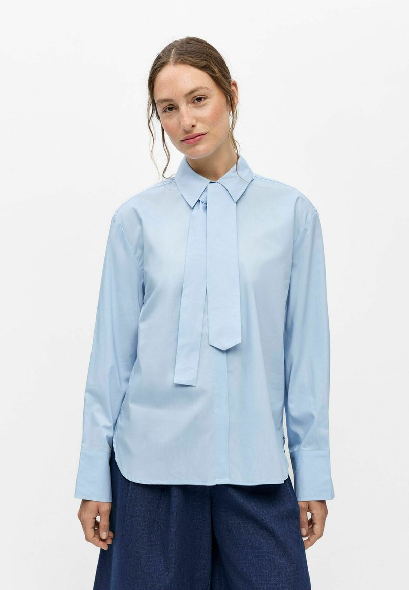 Jeune femme aux cheveux châtain clair porte une chemise bleu clair à manches longues avec un détail de nœud en tissu et un pantalon large bleu foncé.