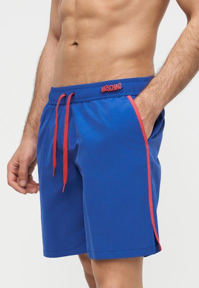 Mann iført blå badeshorts med røde snorer og piping, Moschino-logo på linningen, en hånd i lommen.