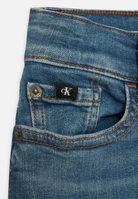 Blå denimjeans med orange söm, med en liten framficka, metallknapp och en svart etikett med "cK"-logotyp.