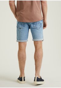 Man som bär ljusblå uppvikta denimshorts, brun t-shirt och marinblå skor med beige sula, står vänd bort på en enkel bakgrund.