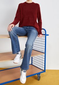 Personne portant un pull rouge, un jean bleu et des baskets blanches assise au bord d'un chariot roulant bleu en métal et en bois.