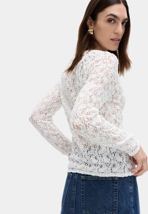 Femme portant un haut en dentelle blanc à manches longues et une jupe en jean bleue, légèrement tournée avec une main sur la hanche, regardant par-dessus son épaule.