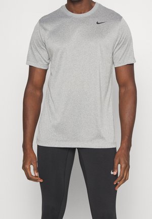 Mann trägt ein hellgraues Nike T-Shirt mit kurzen Ärmeln und schwarze Nike Sport-Leggings, stehend vor weißem Hintergrund.