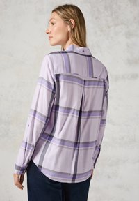 Camisa de cuadros en púrpura claro con mangas largas, detalle de yugo trasero, un solo botón en los puños y dobladillo curvado. La tela parece suave y texturada.