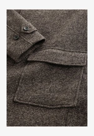 Close-up of brown wool coat sleeve with two black buttons and a flap pocket with textured fabric detail.