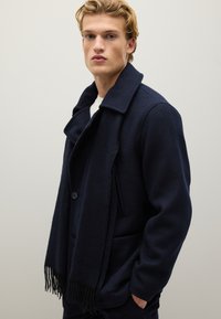 Cappotto in lana blu navy con colletto a revers, chiusura con un solo bottone e tasche laterali. Abbinato a una sciarpa con frange coordinata per un calore aggiuntivo.