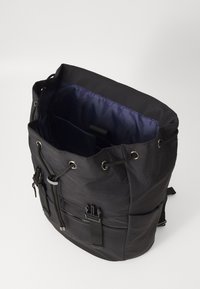 Zaino cilindrico nero con chiusura a cordoncino, dotato di hardware argentato, spallacci regolabili e fodera interna viola. Presenta anche una tasca frontale con zip.