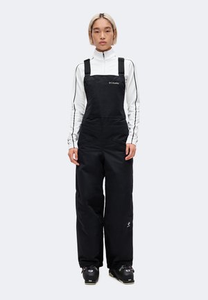 Personne aux cheveux blonds courts portant une combinaison isolante noire, une veste blanche zippée et des chaussures de ski noires, debout face à l'avant.