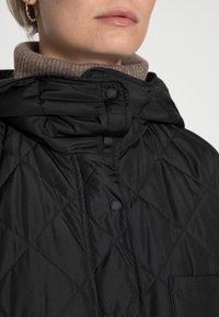 Chaqueta acolchada negra con cuello alto, botones de presión y un bolsillo en la parte delantera. La tela aparenta ser suave con una textura ligera.