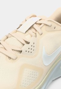 Αθλητικό παπούτσι ανοιχτού μπεζ χρώματος με κορυφή από δίχτυ και συνθετικά υλικά, διαθέτει επίπεδα κορδόνια, διακοσμητικές περφοραρίσεις και λευκό λογότυπο στο πλάι.