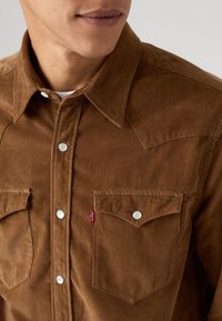Bruine corduroy overhemd met een puntige kraag, twee borstzakken, zilveren drukknoopjes en een kleine rode Levi's-tag op de linkerzak.