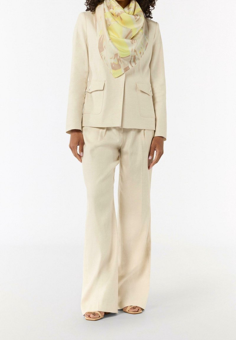 Femme portant un tailleur crème avec un pantalon à jambes larges, un blazer assorti, une écharpe jaune à motifs et des sandales à bouts ouverts.