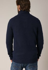 Pull en maille bleu marine avec un col haut, présentant un tissu texturé et des poignets et un ourlet côtelés. Porté avec un jean bleu.