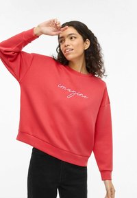 Sudadera roja de manga larga con cuello redondeado. Presenta un texto bordado en blanco que dice "imagine" en la parte delantera. Textura de tela lisa.