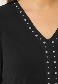 Top nero con scollo a V caratterizzato da borchie decorative argentate lungo il colletto, tessuto morbido e silhouette aderente.
