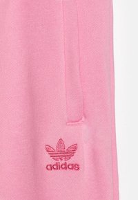 Vaaleanpunaista kangasta, jossa on punaisella brodeerattu Adidas Trefoil -logo ja teksti lähellä taskusaumaa vaatteessa.