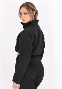 Top corto nero con colletto alto, maniche lunghe e vestibilità morbida. Texture liscia, abbinato a pantaloni neri a vita alta. Design minimale.