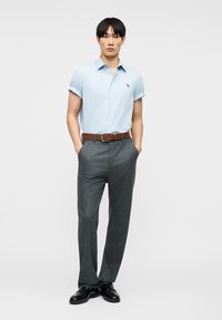 Mann trägt hellblaues, kurzärmeliges Hemd mit Knöpfen, graue, maßgeschneiderte Hose, braunen Gürtel und schwarze Anzugschuhe, steht mit den Händen in den Taschen.