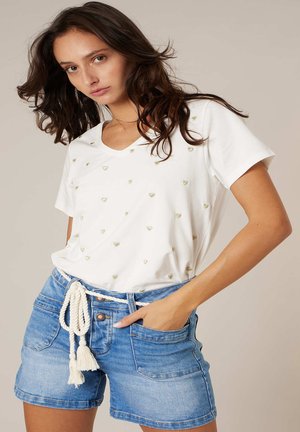 Femme portant un t-shirt blanc à encolure en V avec de petits cœurs dorés et un short en denim bleu avec une ceinture en corde blanche, posant avec une main dans la poche.