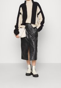 Personne portant un pull rayé noir et beige, une jupe midi en cuir noir boutonnée, des bottines crème et tenant une pochette beige.