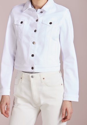 Osoba nosząca białą, krótko skrojoną kurtkę dżinsową zapinaną na guziki na jasnoszarym topie oraz białe, wysokim stanem jeansy na jednolitym tle.