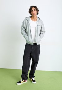 Hanorac gri cu fermoar, având un buzunar tip kangaroo, asociat cu pantaloni negri cu picioare largi și adidași negri cu accente colorate de flacără.