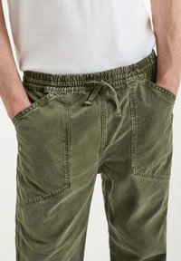 Persoon in olijfgroene drawstring broek met grote voorzakken en een wit shirt, met handen in de zakken.