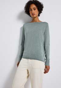 Maglione lavorato a maglia verde chiaro con scollo a barca, maniche lunghe e polsini a coste, abbinato a pantaloni color crema con tasche posteriori.