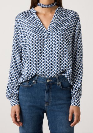 Kobieta w białej bluzce z długim rękawem w niebieskie grochy, zestawiona z niebieskimi dżinsami i pasującą chustą na szyję.