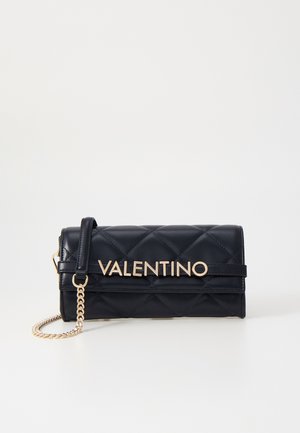 Schwarze gesteppte Valentino Clutch mit goldener Kette und großem goldenem Logo vorne vor weißem Hintergrund.