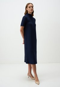 Abito blu navy a maniche corte con colletto alto, texture a costine e taglio dritto, che arriva a metà polpaccio. Indossato con tacchi di colore chiaro.