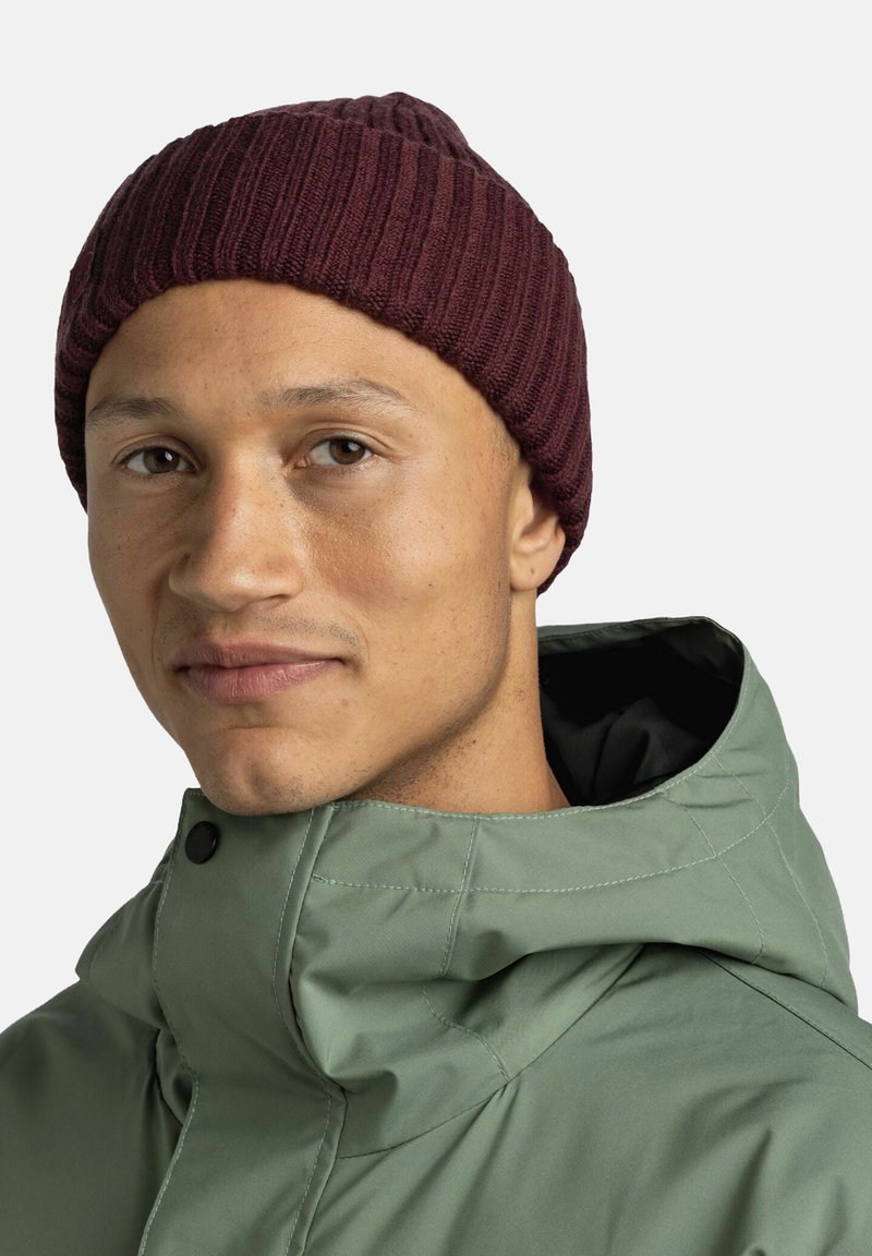 Knit maroon beanie with ribbed texture, worn over the forehead. Green jacket with a high collar and black lining, partially visible.