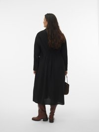 Ženska z dolgimi temnimi lasmi, oblečena v črno midijevo obleko, rjave škornje do kolen, v roki drži majhno rjavo torbico, stoji pred belim ozadjem.