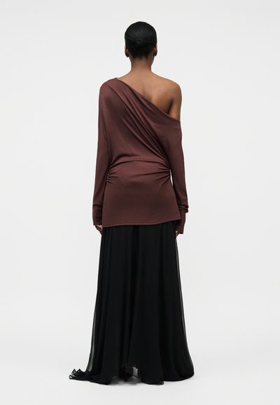 Kobieta z włosami upiętymi w kok, ubrana w brązową, długą bluzkę z odkrytym jednym ramieniem oraz czarną, zwiewną długą spódnicę, stojąca tyłem na jednolitym tle.