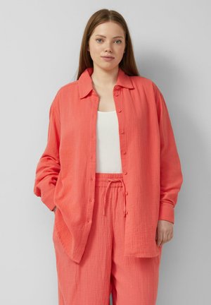 Kobieta ubrana w luźną koralową koszulę zapinaną na guziki oraz dopasowane spodnie ze sznurkiem, na białym topie, stojąca na jednolitym jasnym tle.