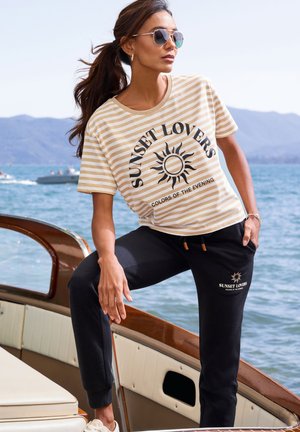 Eine Frau mit Sonnenbrille und gestreiftem "Sunset Lovers"-T-Shirt sowie schwarzer Hose steht auf einem hölzernen Boot am Wasser vor Bergen.
