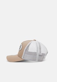 Beige cap with a solid front and mesh back, featuring a round black logo patch on the front. Curved brim with stitched details.