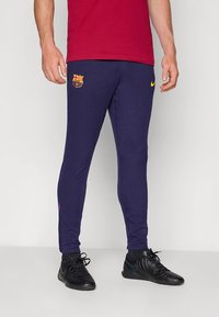 Calças desportivas azul-marinho com textura canelada, apresentando um logótipo do Barcelona e o símbolo Nike em amarelo. Usadas com sapatos desportivos pretos.