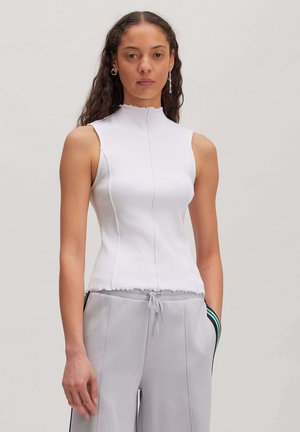 Young woman with long curly hair wearing sleeveless white ribbed top and light gray drawstring pants with side stripes.