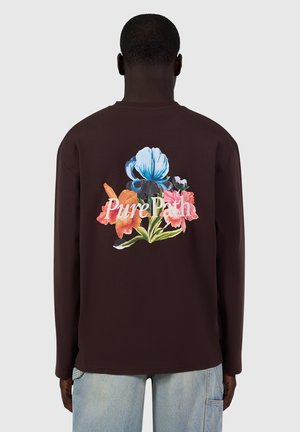 Man wearing a dark brown long-sleeve shirt with colorful flowers and "Pure Path" text printed on the back, paired with light blue jeans.