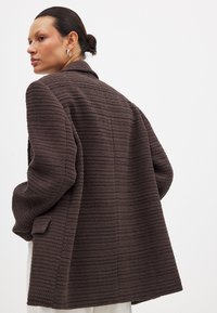 Brauner, strukturierten Blazer mit lockerer Passform, der einen Reverskragen und ein vertikales Streifenmuster aufweist. Die Rückansicht hebt das maßgeschneiderte Design hervor.