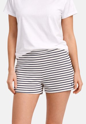 Vrouw draagt een wit T-shirt en witte korte broek met horizontale marineblauwe strepen, staat met ontspannen armen langs het lichaam.