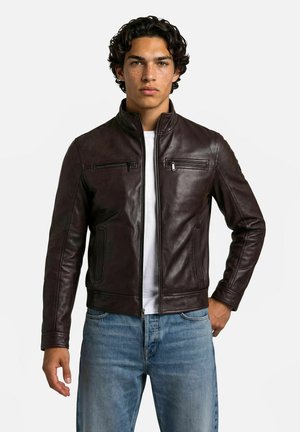 Jeune homme aux cheveux bouclés foncés portant une veste en cuir marron foncé par-dessus une chemise blanche et un jean bleu, debout avec une main sur la hanche.