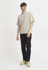 Kortermet beige bomull t-skjorte med et uformelt logo, kombinert med svarte bukser og lyse beige sko, vist på en stående modell.