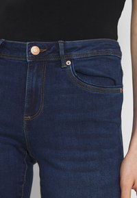 Dark blue denim jeans featuring a high waist, copper button, and gold stitching details on pockets and seams.