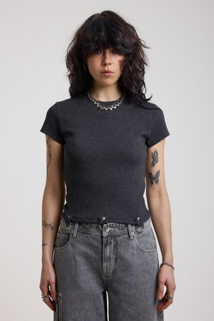 Γυναίκα με σκούρα σγουρά μαλλιά που φοράει εφαρμοστή σκουρόγρια κοντομάνικη μπλούζα, γκρι τζιν, ασημένιο κολιέ και εμφανή τατουάζ στα χέρια, στέκεται μπροστά σε λευκό φόντο.