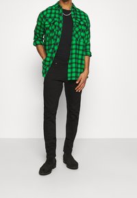 Man med en grön och svart rutig skjorta över en svart t-shirt, svarta byxor, svarta stövlar och silverkedja hängande runt halsen, står med händerna i fickorna.